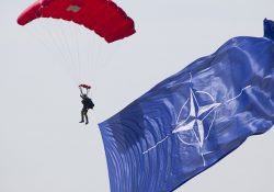 a parachuter in the air with a large NATO flag behind them.